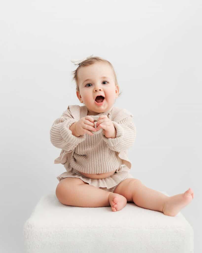 Image of one year old baby sitting on white ottoman. White background. Baby is wearing tan sweater and tan shorts.
