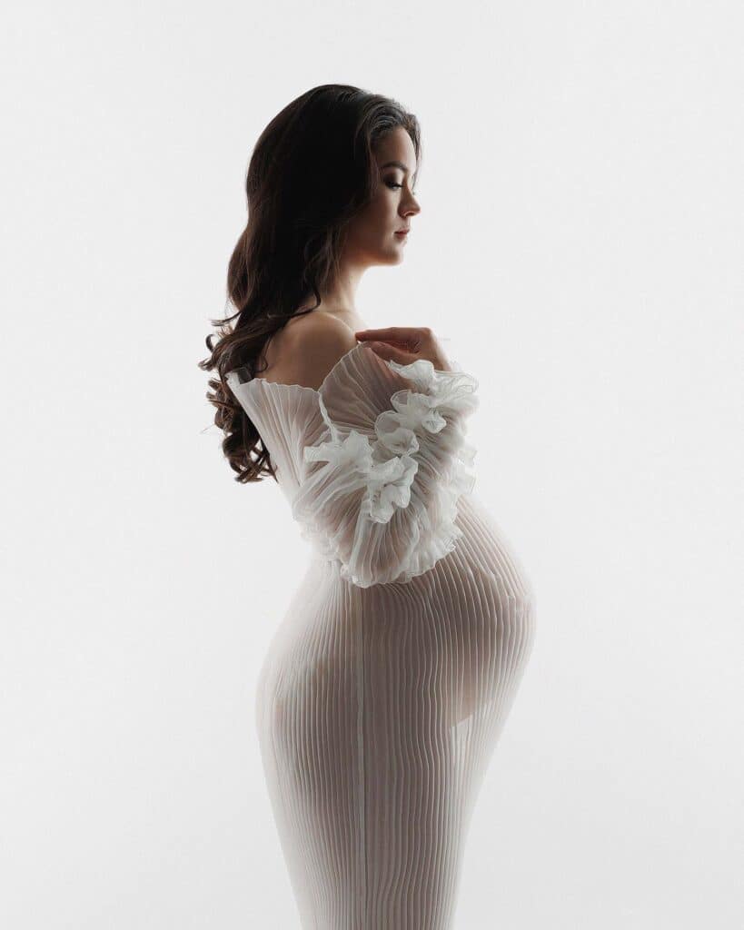 Image of women to sheer white dress, with a white backdrop. Women is pregnant.