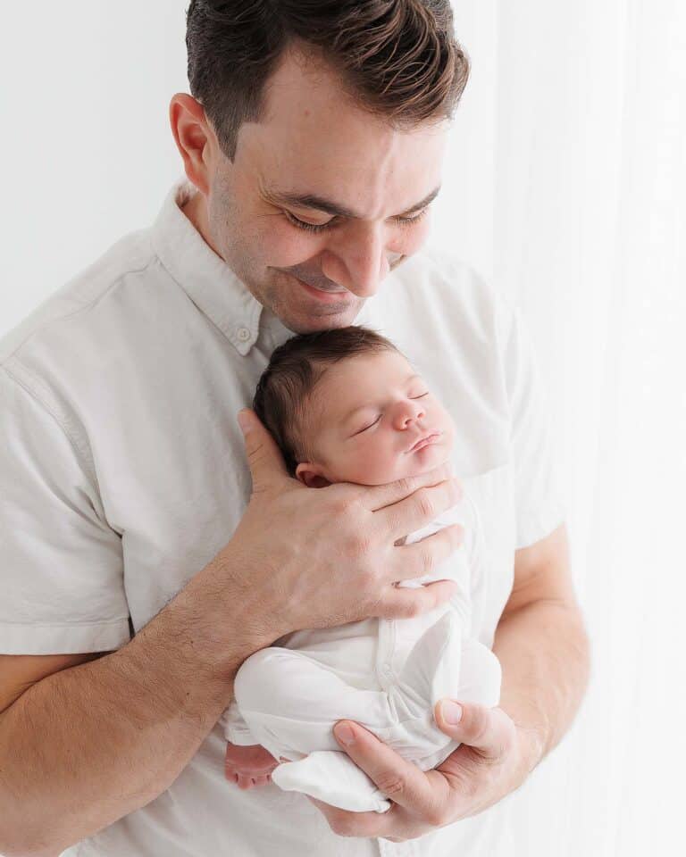 Image of man smiling, holding his newborn baby. Man is wearing white, short sleave dress shirt and baby is in white, fitted pajamas.