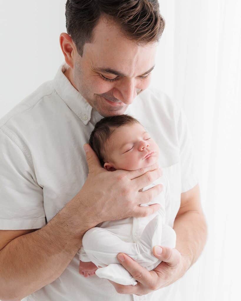 Image of man smiling, holding his newborn baby. Man is wearing white, short sleave dress shirt and baby is in white, fitted pajamas.